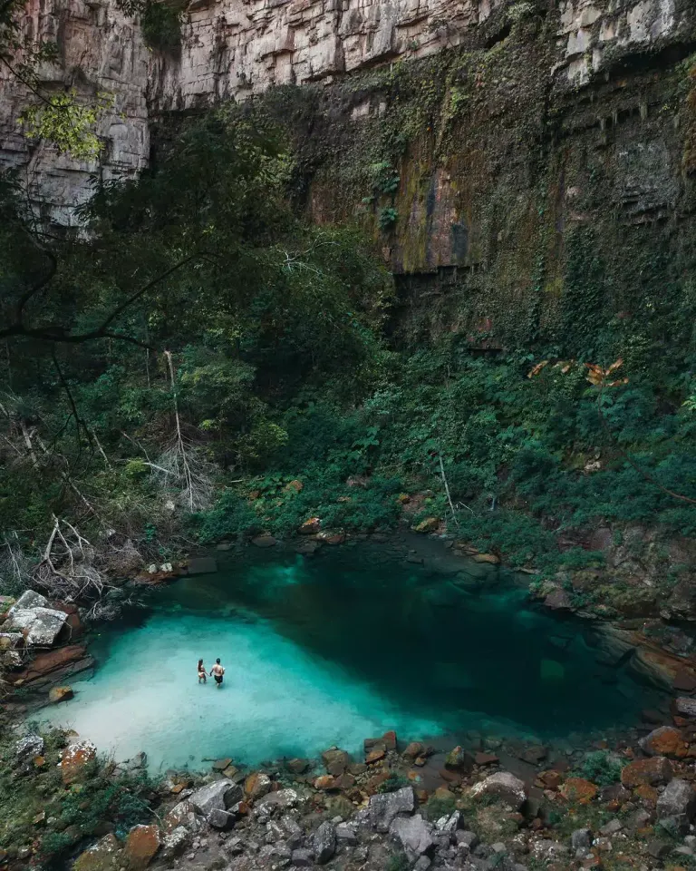 Vista de cima do Santuário das Araras, poço de água verde-azulada ao pé de um paredão de pedra, com duas pessoas entrando na água