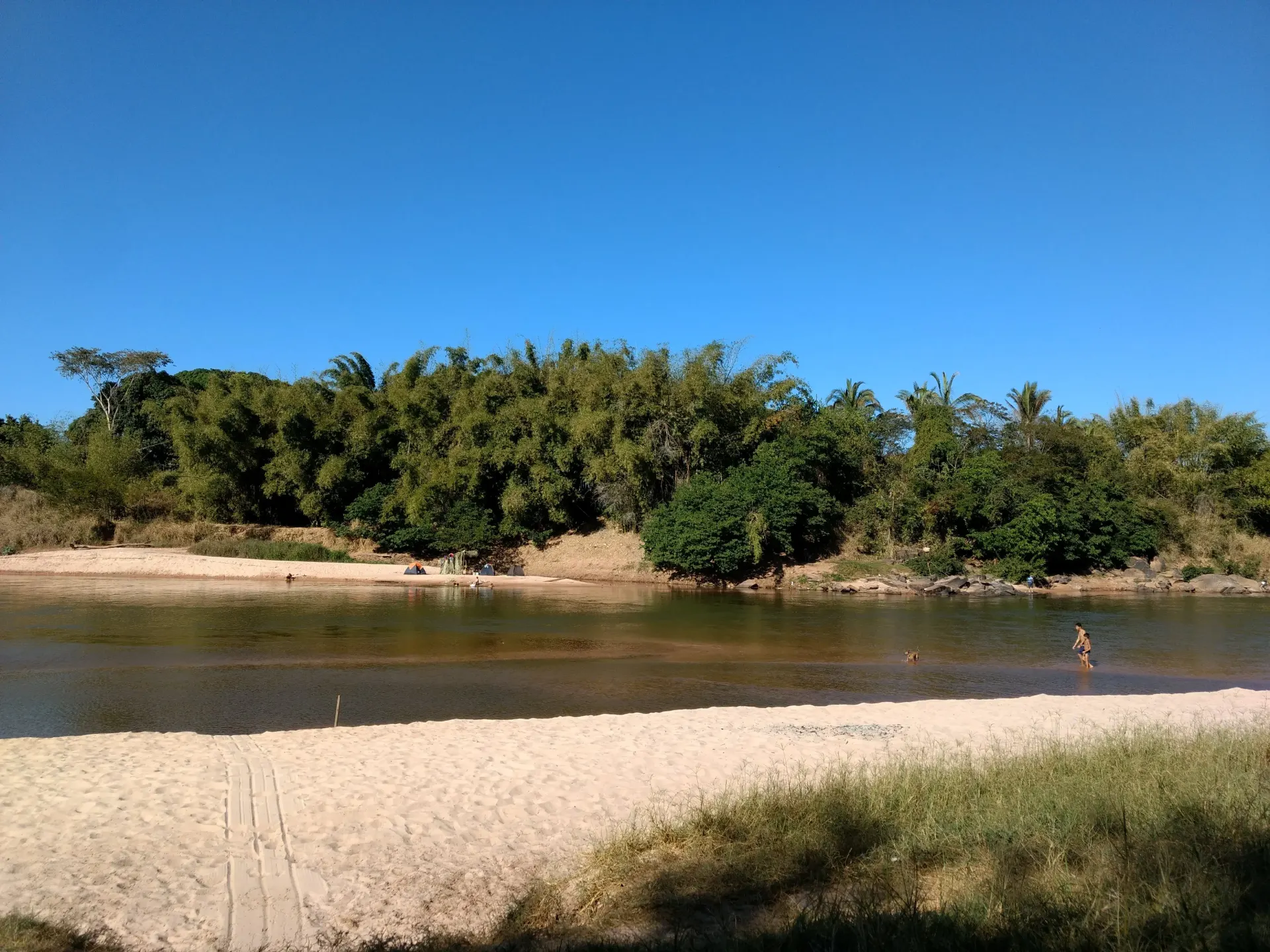 foto que mostra uma pequena parte da praia, tem algumas pessoas na água e do outro lado do rio tem a beirada cheia de arvores.
