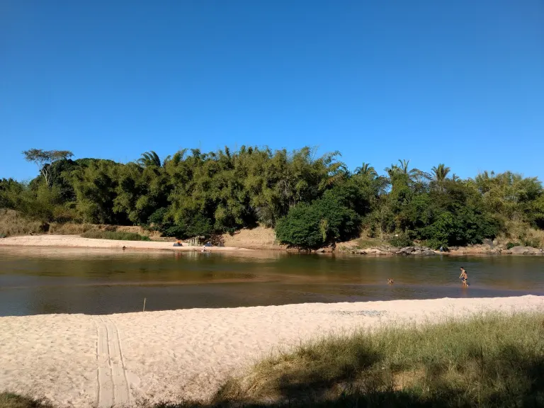 foto que mostra uma pequena parte da praia, tem algumas pessoas na água e do outro lado do rio tem a beirada cheia de arvores.